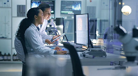Two people seated in a lab workstation comtemplate and discuss what they see on a large computer monitor